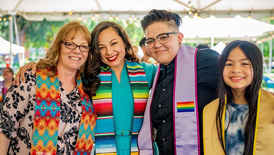 A group of women and one girl under a big event tent and leaning into each other wearing graduation stoles representing Native American, Hispanic, Queer, and AAPI identities
